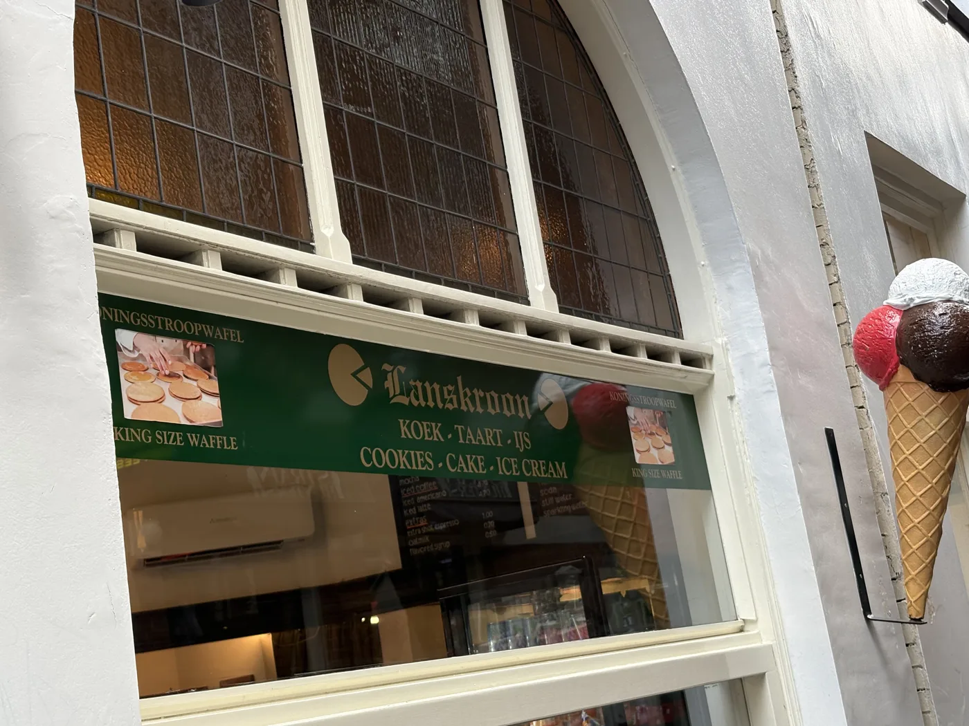 Etalage van Lanskroon met het groene opschrift Koek, Taart, IJs en foto's van de Koningsstroopwafel onder het glas-in-loodraam