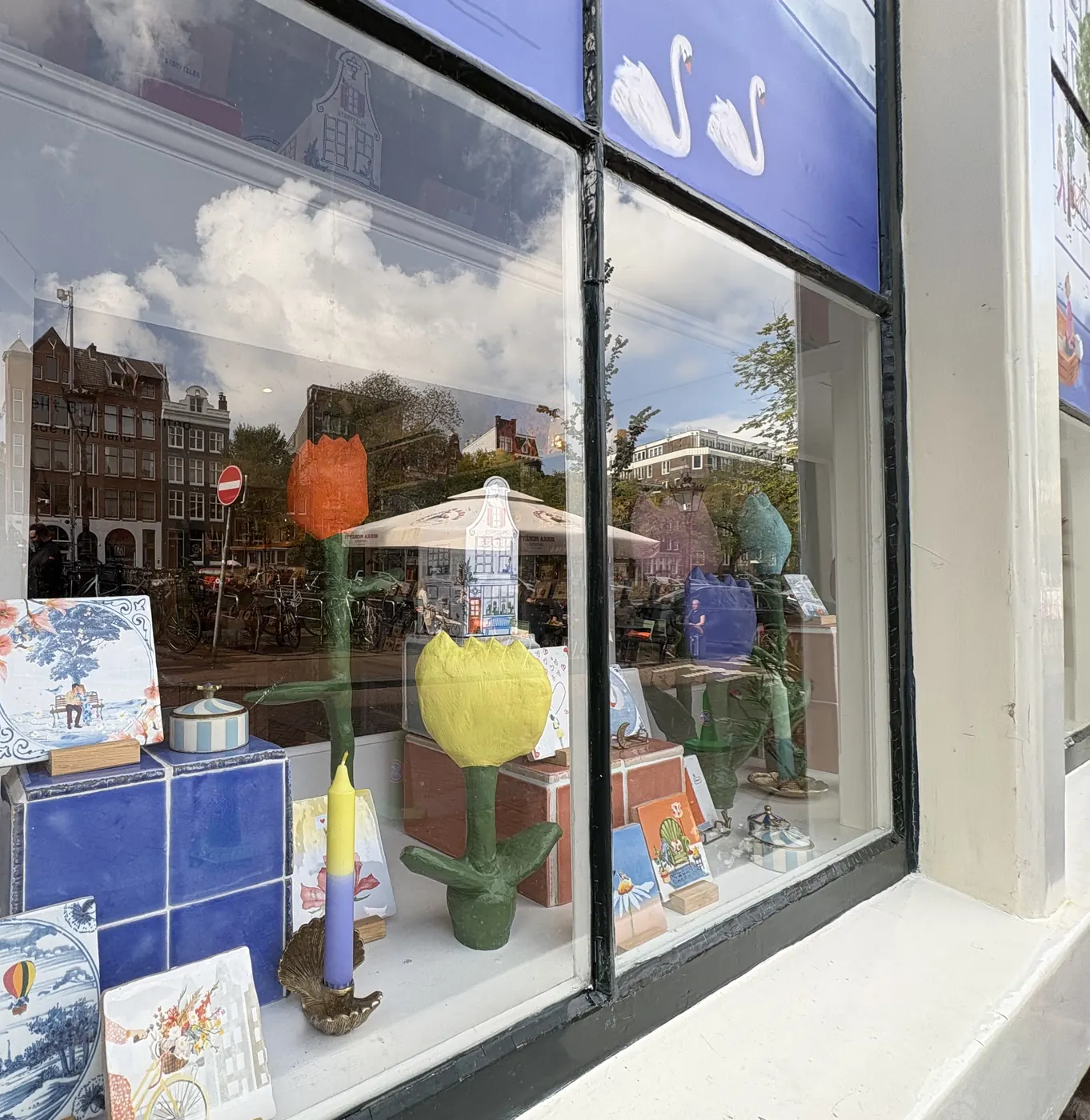 Etalage van StoryTiles aan het Singel met grote gekleurde tulpen, een kaars en tegels op houten standaardjes, met in het glas de spiegeling van de overkant