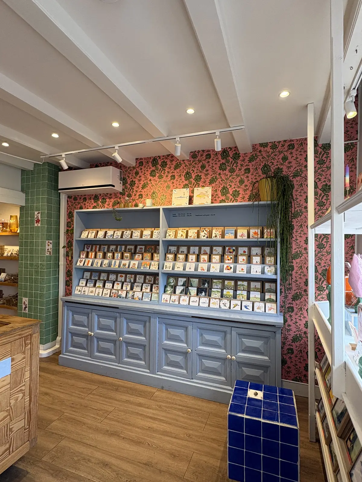 Lichtblauwe kast vol tegels tegen roze bloemetjesbehang in de winkel van StoryTiles, met een blauw betegeld krukje ervoor